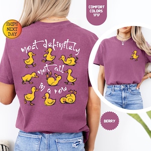 May include: Berry-colored t-shirt with a playful design featuring yellow cartoon ducks and the phrase "most definitely not all in a row." A small duck graphic is on the front. The shirt is a Comfort Colors 1717.