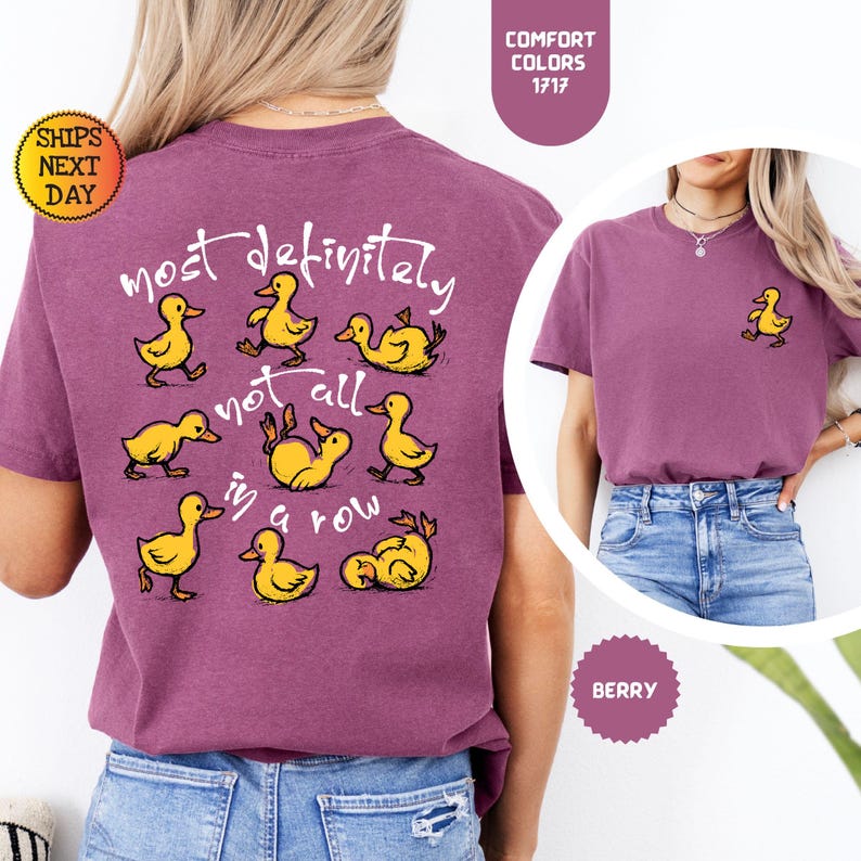 May include: Berry-colored t-shirt with a playful design featuring yellow cartoon ducks and the phrase "most definitely not all in a row." A small duck graphic is on the front. The shirt is a Comfort Colors 1717.