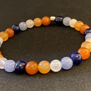 Puede incluir: Un brazalete hecho de cuentas azules, naranjas y blancas. Las cuentas están enhebradas en un cordón plateado.