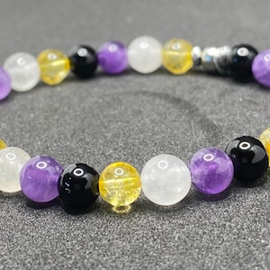 Op de afbeelding: Een armband gemaakt met amethist, citrien, witte jade en zwarte onyx kralen. De kralen zijn gerangschikt in een patroon dat lijkt op de kleuren van de non-binaire pride-vlag.