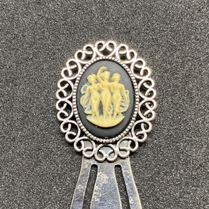 May include: Silver bookmark with an oval cameo depicting three cream-coloured figures on a black background. The cameo is set within a decorative silver filigree frame. The bookmark has a long, slender design.