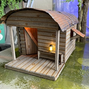Puede incluir: Una cabaña de madera en miniatura con un techo de metal oxidado, un porche y una pequeña linterna en el porche. La cabaña está sentada sobre una superficie verde.