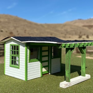 Puede incluir: Maqueta de cobertizo blanco y verde con techo negro. El cobertizo tiene una pequeña ventana y un porche cubierto sostenido por postes verdes. La estructura está sobre una superficie verde, con un fondo borroso de colinas.
