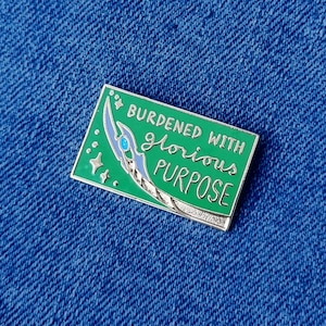 Op de afbeelding: Een rechthoekige emaille pin met een groene achtergrond en de tekst "BURDENED WITH GLORIOUS PURPOSE". De pin heeft een zilver en blauw ontwerp. De pin is geplaatst tegen een blauwe denim achtergrond.