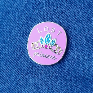 May include: A pink enamel pin with a silver crown and the words "LOST princess" in white.