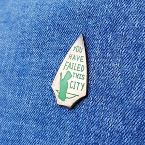 May include: A silver arrowhead-shaped pin with a green silhouette of a hooded figure and the text "You Have Failed This City".