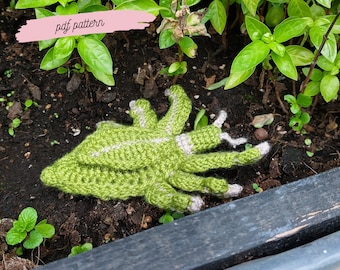 Op de afbeelding: Een groen en wit gehäkeld alien-wezen met meerdere armen en benen. Het wezen ligt op de grond in een tuinomgeving. De tekst "pdf pattern" staat op een roze banner boven het wezen. De tekst "rainyfrogs.etsy.com" staat op het lichaam van het wezen.