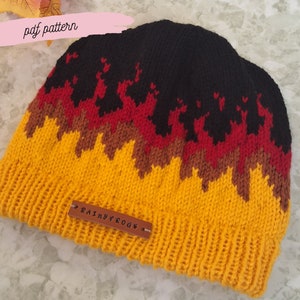 Puede incluir: Un gorro de punto negro, amarillo, rojo y marrón con un patrón de llamas. El gorro tiene una etiqueta de cuero que dice "Rainy Frogs".