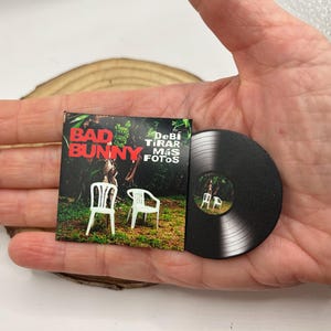May include: A miniature vinyl record of Bad Bunny's album "Un Verano Sin Ti" with the album cover art featuring two white plastic chairs in a tropical setting. The album cover text reads "Debi Tirar Mas Fotos".