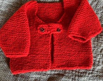 May include: A bright red hand-knitted cardigan featuring a garter stitch texture, long sleeves, and a round neckline. The front of the garment is secured with two dark brown buttons positioned at the center of the chest area, creating a simple closure.