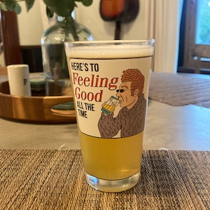 May include: A clear glass with a white and yellow beverage. The glass has a cartoon illustration of a person with red hair wearing a sweater and sunglasses, drinking from a glass. The text on the glass reads "Here's to Feeling Good All the Time."