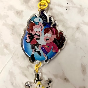 May include: A clear acrylic charm featuring characters from the animated series Gravity Falls. The charm has a yellow star-shaped top, a central image of two characters in a blue flame design, and a yellow triangle character at the bottom.