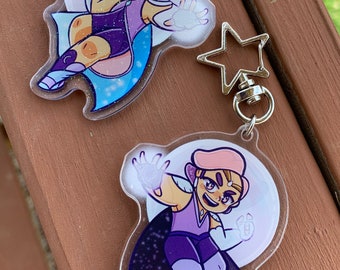Op de afbeelding: Twee doorzichtige acryl sleutelhangers met glitter ingebed in het plastic. Elke sleutelhanger heeft een cartoonfiguur met roze haar, een paarse outfit en een witte cape. Het figuur links staat op een halve maan met een blauwe en witte sterrenhemel op de achtergrond. Het figuur rechts staat op een paarse en zwarte sterrenhemel op de achtergrond.