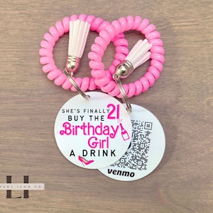 May include: Two pink spiral hair ties with white tassels attached to a silver key ring with two round white tags. One tag says "She's finally 21 Buy the Birthday Girl a Drink" with a pink high heel shoe icon. The other tag has a QR code and the word "venmo" printed on it.
