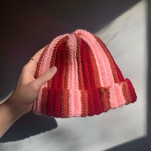 May include: A handmade, striped beanie in shades of pink, red, and brown. The ribbed knit hat features a folded brim and is held by a hand. The color scheme creates a warm, cozy aesthetic, perfect for cold weather.