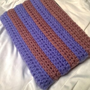 May include: A crocheted striped pillow cover in shades of purple and brown. The cover is made with a textured stitch and has a rectangular shape.