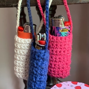 May include: Three crocheted zipper pouches in white, blue, and pink, each holding a lighter. The lighters are all different designs. The pouches are hanging from a wooden shelf.