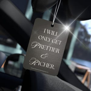 Puede incluir: Un ambientador rectangular gris oscuro para coche con el texto "I WILL ONLY GET PRETTIER & RICHER" en escritura blanca. El ambientador está colgado por un hilo blanco, con una luz brillante en el fondo.