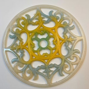 May include: A round, white, decorative coaster with a raised, intricate design. The design is a repeating pattern of swirls and loops, with a central flower shape. The coaster is painted in shades of yellow, green, and blue.