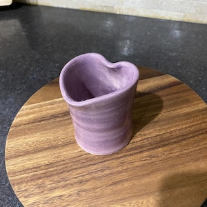 May include: A handmade, lavender-colored ceramic heart-shaped container. The container is cylindrical with a heart-shaped opening at the top. It sits on a round, wooden serving board. The background is a dark countertop and a tiled backsplash.