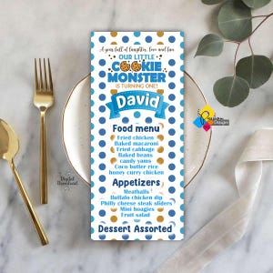 May include: A blue and white polka-dotted menu card with the text "Our Little Cookie Monster is Turning One!" and "David." The menu lists food items like fried chicken and mini hoagies. Gold cutlery and a plate are in the background.