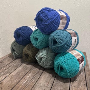 May include: A collection of colorful yarn balls in shades of blue, green, and olive arranged on a wooden surface. Each ball has a label wrapped around it. The yarn appears to be a thick, textured material, suitable for various crafting projects.