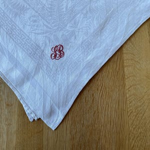 May include: White linen handkerchief with a floral pattern and a red embroidered monogram. The handkerchief is folded, displaying the corner with the monogram. The fabric has a subtle texture, and the monogram is in a script style.