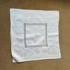 Può includere: Tovaglia bianca con motivo floreale e bordo decorativo. Il bordo quadrato presenta un motivo geometrico ripetuto. La tovaglia è realizzata in materiale morbido, possibilmente lino.