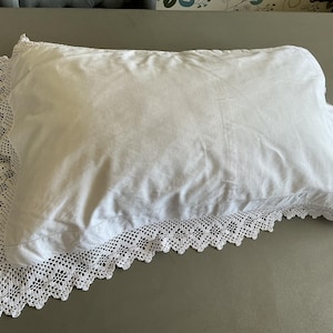 Puede incluir: Una funda de almohada rectangular blanca con un borde decorativo de encaje de ganchillo blanco a lo largo de los lados. La tela es lisa y sencilla, contrastando con el intrincado patrón calado del delicado encaje que enmarca la almohada.