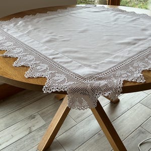 May include: A square white fabric tablecloth featuring a smooth center panel bordered by an intricate, wide crocheted lace edge with a pointed, scalloped design. The lace trim displays a detailed floral pattern throughout the entire perimeter.