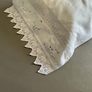 Może przedstawiać: Biała poszewka na poduszkę z misternymi koronkowymi detalami na krawędzi. Koronka ma kwiatowe i motylkowe wycięcia, dodając delikatnego charakteru. Poszewka wykonana jest z miękkiej, gładkiej tkaniny, idealnej do pościeli.