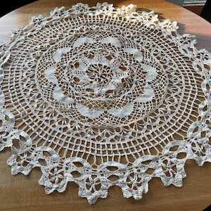 Könnte beinhalten: Cremefarbene, runde Häkeldecke mit detailliertem Lochmuster. Die Decke hat ein zentrales Blumenmuster und einen gewellten Rand. Die filigrane, spitzenartige Textur ist auf der Holzoberfläche sichtbar.