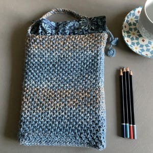May include: A blue and brown crocheted bag with a fabric lining and a handle. The bag is sitting on a gray surface with four pencils next to it.