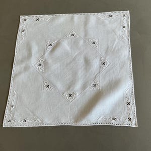 Puede incluir: Una servilleta cuadrada de lino blanco con intrincados bordados calados. El diseño incluye un borde central en forma de diamante con motivos florales en las esquinas y a lo largo de los bordes, acabado con un delicado detalle de vainica.