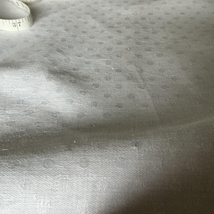 May include: A white fabric featuring a subtle, repeating tonal dot pattern across its surface. A white measuring tape is visible in the upper left corner, showing markings for 36 and 37 inches, providing a clear scale for the textile texture.