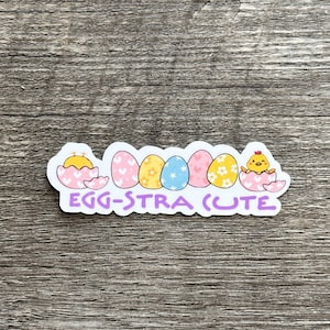 May include: A white sticker with a yellow chick hatching from an egg, surrounded by five colorful eggs with different patterns. The text "EGG-STRA CUTE" is written in purple below the eggs.