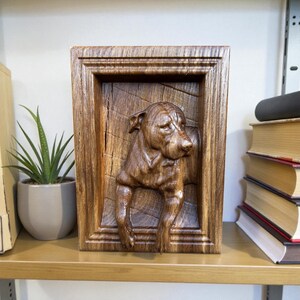 May include: A wooden relief carving of a dog in a rectangular frame. The dog is carved in a realistic style, with detailed fur and facial features. The frame is made of a dark wood with a natural grain. The artwork is displayed on a wooden shelf.