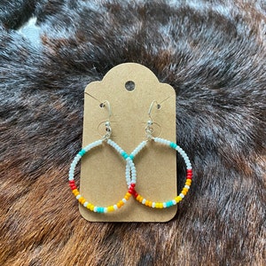 Peut inclure: Boucles d'oreilles créoles avec un motif de perles. Les boucles d'oreilles sont dotées de crochets argentés et de cerceaux circulaires ornés de petites perles colorées blanches, turquoise, jaunes, oranges et rouges. Elles sont présentées sur une carte marron.