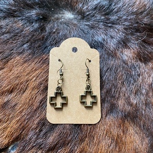 Peut inclure: Une paire de boucles d'oreilles en forme de croix de couleur bronze, présentées sur une carte marron. Les boucles d'oreilles ont un design de croix ouvert et sont suspendues à de petits crochets. Le fond est une surface texturée marron et noire.