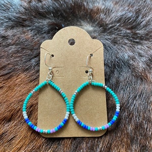 Peut inclure: Boucles d'oreilles en forme de goutte d'eau ornées de perles turquoise, bleues, blanches et argentées. Les boucles d'oreilles sont présentées sur une carte marron avec un petit trou en haut. Le fond est une surface texturée marron et noire.