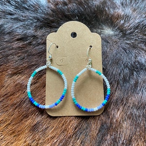 Peut inclure: Boucles d'oreilles créoles ornées de perles. Les boucles présentent un dégradé de couleurs, notamment des perles blanches, turquoise, bleues et lavande. Elles sont suspendues à des crochets argentés et présentées sur une carte marron.