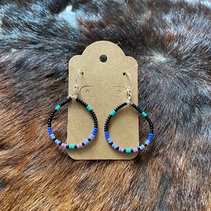 Peut inclure: Boucles d'oreilles créoles avec un motif de perles. Les boucles d'oreilles ont des crochets argentés et sont ornées de petites perles colorées noires, bleues, turquoise et lavande. Elles sont présentées sur une étiquette marron sur un fond texturé marron et noir.