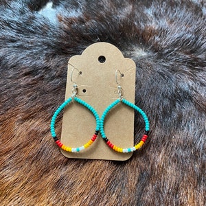Peut inclure: Boucles d'oreilles créoles en perles turquoises avec un motif coloré de perles rouges, jaunes, noires et blanches. Les boucles d'oreilles sont suspendues à des crochets argentés et sont présentées sur une carte marron.