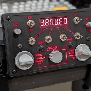 May include: A black radio control panel featuring a red digital display showing 225.000. The device includes multiple silver toggle switches, three grey rotary knobs, and red printed labels such as VOL, SQL, TONE, LOAD, CHAN SEL, GUARD, MANUAL, PRESET, ADF, BOTH, MAIN, OFF, AD, and BRT.