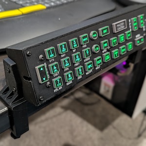 May include: Black control panel with green illuminated buttons and text. The panel is mounted on a desk with a clamp. The buttons are labelled with numbers, letters, and function names. The text "MASTER CAUTION" is visible.