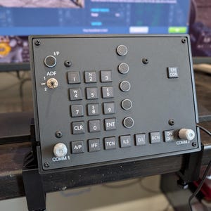 May include: A black rectangular flight simulator control panel featuring a numeric keypad, a toggle switch labelled ADF, and various buttons labelled I/P, EM CON, A/P, IFF, TCN, ILS, D/L, BCN, and ON OFF. Two silver knobs labelled COMM 1 and COMM 2 are present.