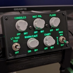 Puede incluir: Un panel de control negro con múltiples perillas e interruptores. El panel tiene etiquetas para "Consoles", "Inst Pnl", "Flood", "Lt Test", "Warn/Caut", "Chart", "Mode NVG", "Dim" y "Nite/Day".