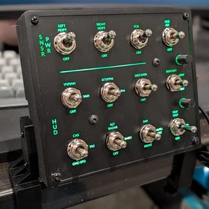 May include: A black control panel with multiple toggle switches and green illuminated text. The panel includes labels such as "SNSR", "PWR", "HUD", and various function indicators. It is mounted on a black stand.