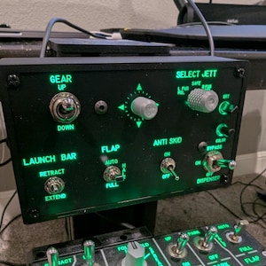 May include: A black control panel with green illuminated buttons and labels. The panel features controls for gear, launch bar, flaps, anti-skid, and select jett. The labels include "Gear Up", "Gear Down", "Launch Bar Retract", "Launch Bar Extend", "Flaps Auto", "Flaps Full", "Anti Skid On", "Anti Skid Off", "Select Jett Safe", "Select Jett L Fus Hol", "Select Jett Bot", "Color Bypass", "Dispenser On", "Dispenser Off".