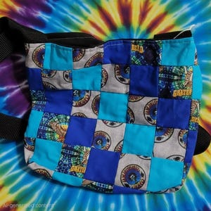 Op de afbeelding: Een patchwork schoudertas met een dambordpatroon van effen blauwe, fel turquoise en grijze stoffen vierkanten. De grijze vierkanten tonen cirkelvormige afbeeldingen met schedels en de tekst Dead, terwijl andere vierkanten tie-dye patronen hebben.